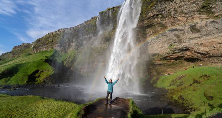 A lone traveler standing with arms raised in front of a tall waterfall and rainbow on a bright day.