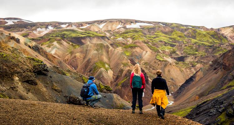 Three hikers pause on a gravel ridge overlooking colourful volcanic valleys and distant snow patches.