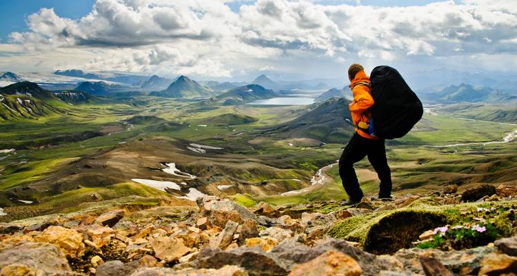 A backpacker stands on a rocky slope overlooking vast green highlands, lakes and distant peaks under dramatic clouds.