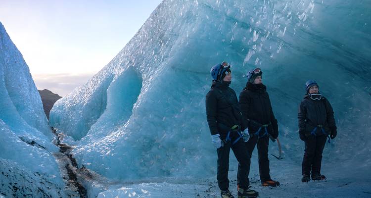Three climbers wearing helmets and crampons explore blue ice walls inside a glacier.