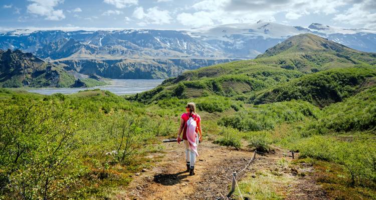 A hiker in pink walks a green ridge with snow-capped peaks and river valley beyond.