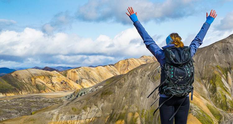 A triumphant hiker raises arms atop colorful striped mountains and valleys.