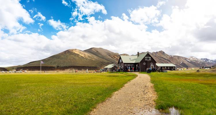 A broad meadow and path lead to a mountain lodge beneath blue skies and patchy clouds.