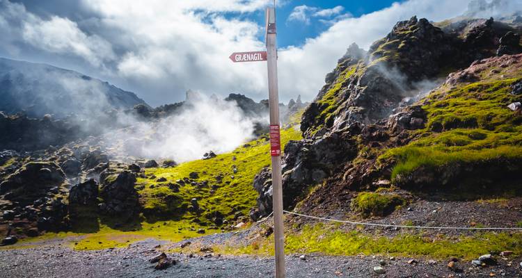 Steam rises from rocky lava fields near a trail sign under dramatic clouds.