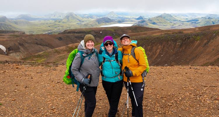 Tres excursionistas sonrientes posando con bastones en una meseta azotada por el viento con vistas a lagos y montañas islandeses