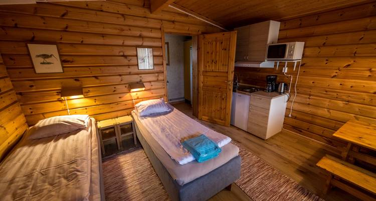 Interior de una acogedora cabaña de madera con camas, cocina pequeña y una ventana.