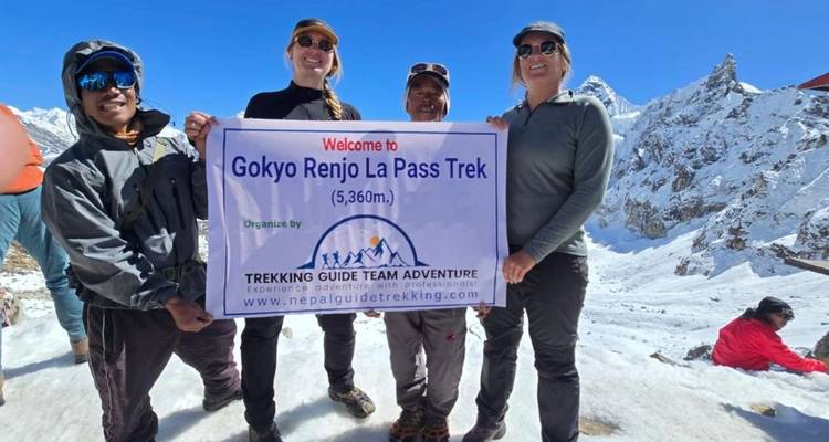 Trekkers sonrientes sosteniendo un banner en el nevado Renjo La Pass con brillantes picos del Himalaya de fondo.