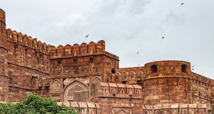 A historic red fort with flying birds and cloudy sky.