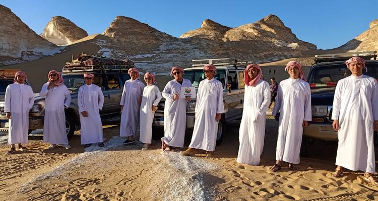 Gruppe von Menschen in traditioneller Kleidung, die vor Geländewagen in einem Wüstengebiet stehen.