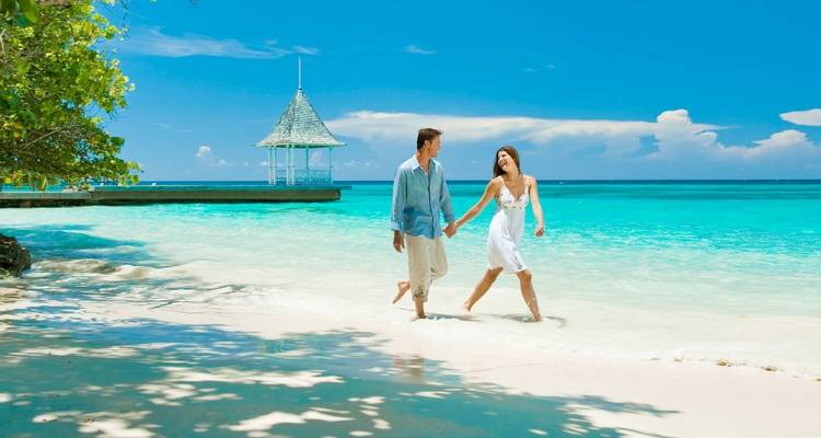Couple walking on a white sandy beach with turquoise water and a gazebo.