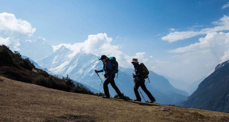 Dos excursionistas en un sendero de montaña con una cordillera nevada al fondo.
