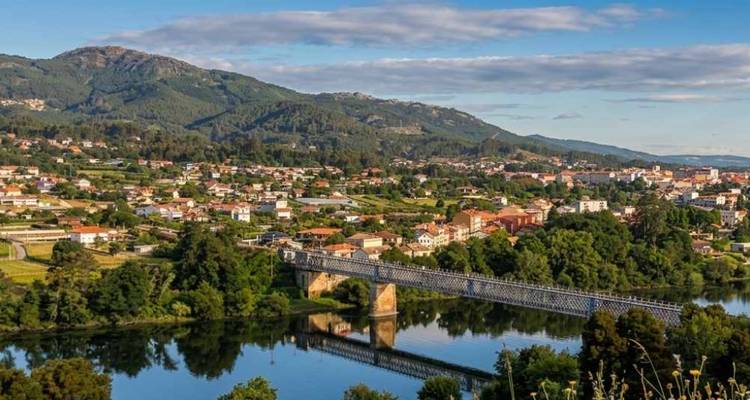 Vallée verdoyante dans le nord du Portugal avec un pont métallique enjambant une rivière aux eaux calmes, dominée par des collines luxuriantes sous un ciel partiellement nuageux.