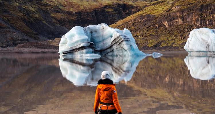 Un voyageur en veste orange contemple un lagon glaciaire réfléchissant avec des icebergs flottants.
