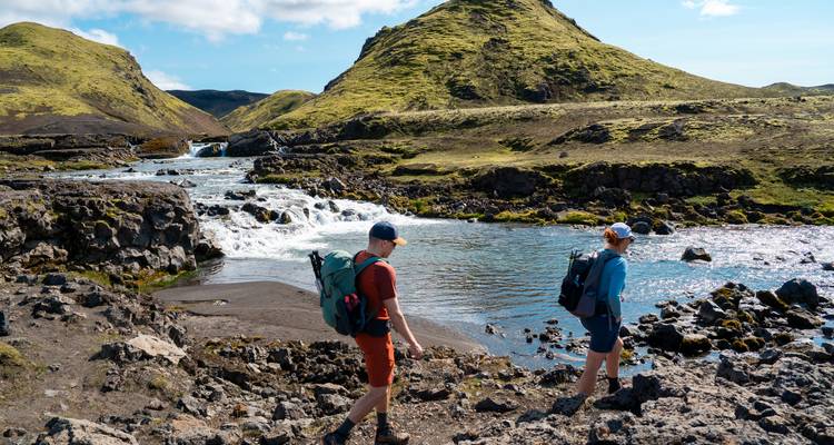 Deux randonneurs explorent les rives rocheuses d'une rivière au-dessous de collines d'un vert éclatant par une belle journée ensoleillée