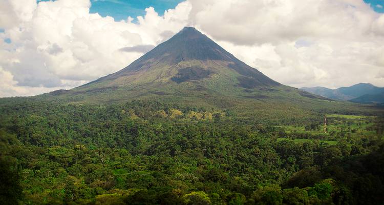 A majestic conical volcano rising above dense tropical forest under a bright blue sky with scattered clouds.