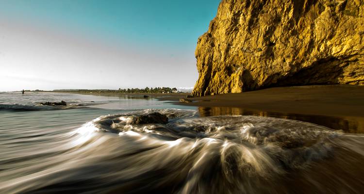 Long-exposure image of waves swirling against a golden seaside cliff at a tranquil beach.