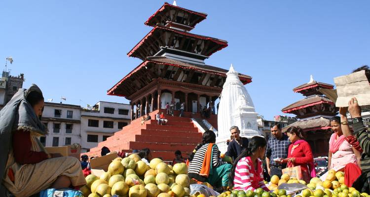 Scène de marché avec des gens vendant des fruits devant un temple à l'architecture complexe.