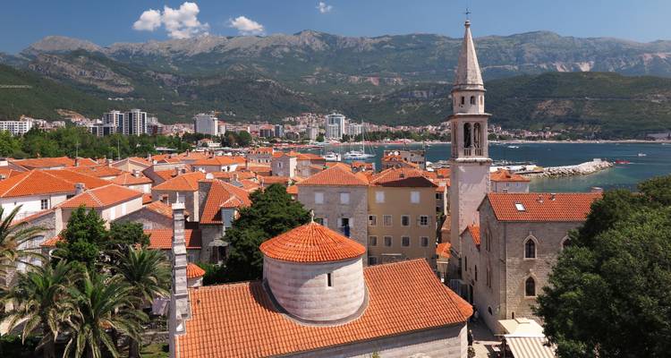 Coastal view with red-roofed buildings, a tall church tower, and mountains in the background.