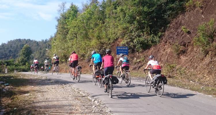 Group of cyclists riding on a paved road with a mountainside view.