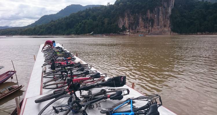 Bicycles on a boat by a river with rocky cliffs in the background.