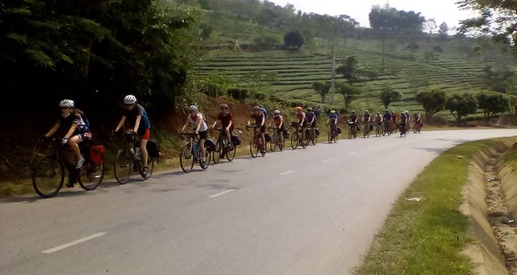 Cyclists riding in a line along a rural road with hills in the background.