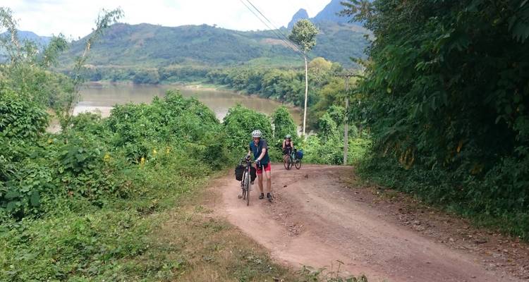 Cyclist riding on a dirt path near a river with foliage.
