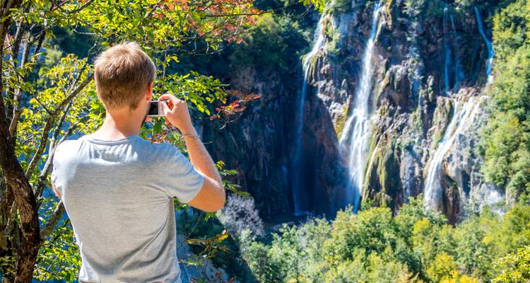 Un hombre fotografía una cascada alta rodeada de follaje otoñal colorido en Plitvice.