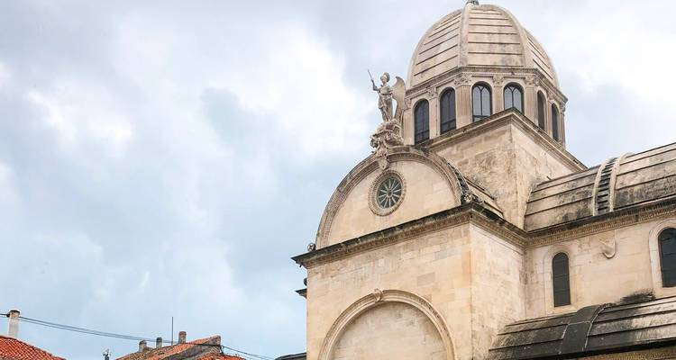 Cúpula y estatua en lo alto de la Catedral de Sibenik enmarcadas contra un cielo nublado y tejados de terracota.
