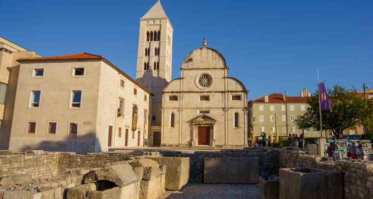 Una iglesia de piedra histórica y su campanario se elevan sobre ruinas romanas antiguas bajo la luz de la tarde en Zadar.