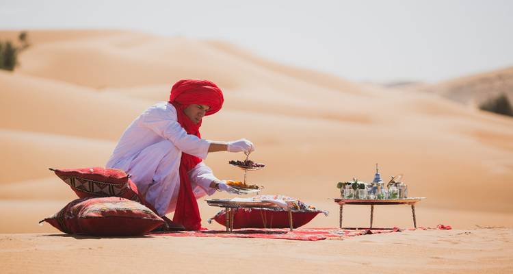 Una persona sentada entre cojines en un entorno desértico con una preparación tradicional de té.