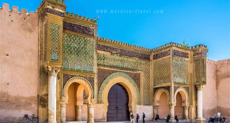 Puerta histórica con trabajo de azulejos intrincado, algunas personas y vehículos al frente.