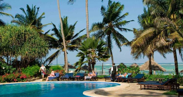 Resort setting with palm trees, a pool, and people relaxing by the beach.