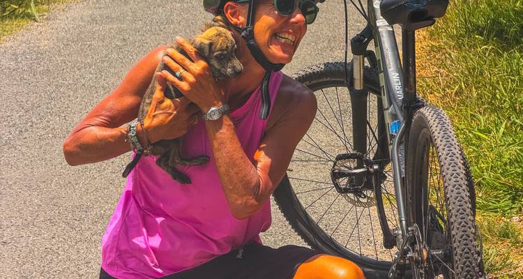Cycliste souriante en maillot rose câline un petit chiot à côté de son VTT sur une route rurale.