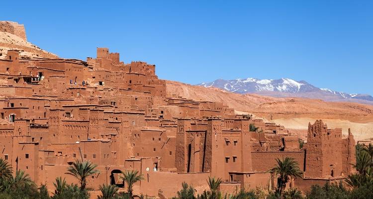 Lehmziegels-Kasbah von Ait Ben Haddou vor einer Kulisse aus trockenen Hügeln und schneebedeckten Atlas-Bergen.