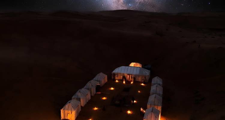 Un campement du désert illuminé avec des tentes blanches disposées en rangées sous un ciel nocturne brillant rempli d'étoiles, entouré de dunes de sable sombre.