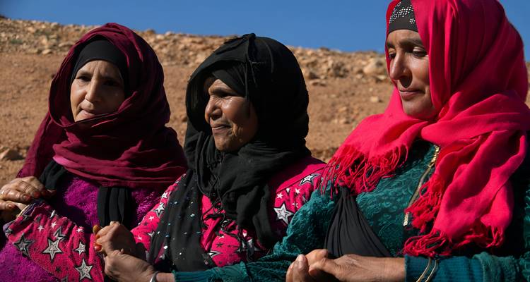 Trois femmes berbères en costume traditionnel vibrant se tenant la main avec un paysage désertique en arrière-plan.