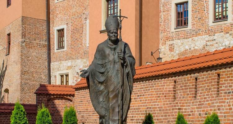 Statue d'une figure religieuse en robes tenant un bâton, positionnée devant un bâtiment historique en briques.