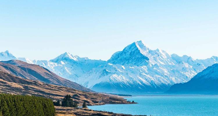 Los Alpes del Sur cubiertos de nieve se alzan sobre un lago turquesa y llanuras ondulantes de pastizales bajo una luz nítida.