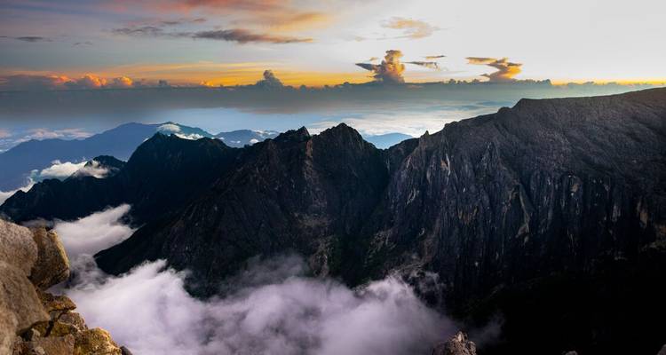 Panoramic vista of Kinabalu’s jagged peaks protruding above clouds with colorful sunrise sky.