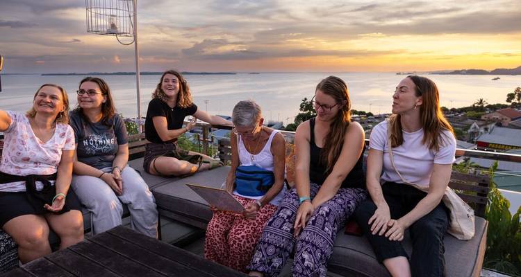 Group of travelers relaxing on a rooftop terrace overlooking the sea during a colorful sunset.