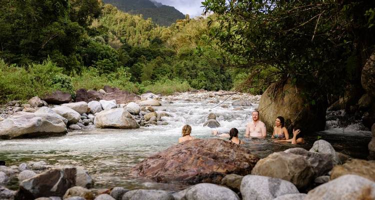 Travelers bathing in a clear rocky river surrounded by tropical forest and mountains.