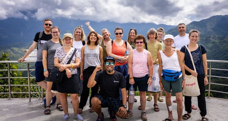 Large tour group smiling at a scenic mountain viewpoint with cloud-covered peaks behind.
