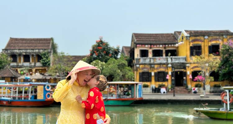 Eine Frau und ein Kind lächeln in traditioneller Kleidung mit bunten historischen Gebäuden im Hintergrund.