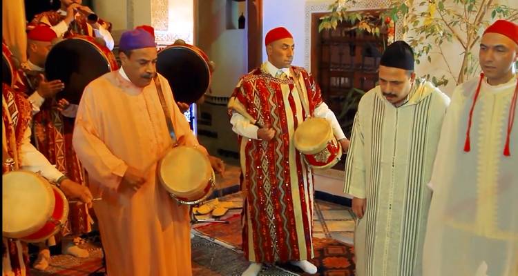 Groep Marokkaanse muzikanten in kleurrijke traditionele kleding bespelen handtrommels in een versierde kamer.