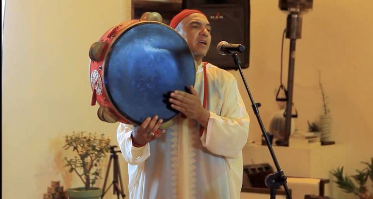 Man in traditionele Marokkaanse gewaad en rode muts treedt op met een grote raamtrommel voor een microfoon binnenshuis.