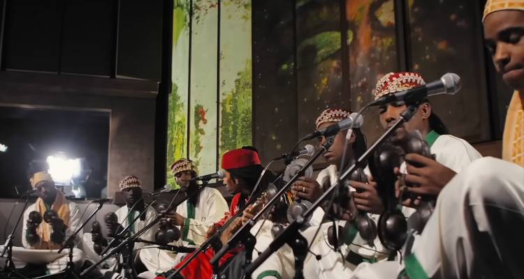 Ensemble van muzikanten in traditionele kledij treedt op met castagnetten en drums op een zwak verlicht podium.