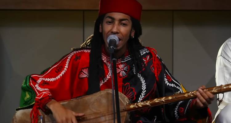 Muzikant in rode fez en geborduurd gewaad zingt in een microfoon terwijl hij een guembri bespeelt.
