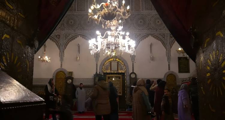 Zwak verlichte binnenkant van een heiligdom of moskee met gelovigen onder sierlijke kroonluchters.