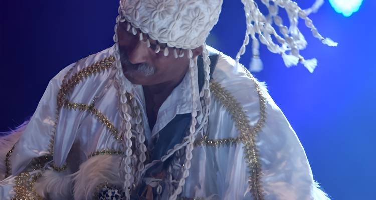 Artiste traditionnel marocain en costume blanc orné et coiffe, saisi en pleine danse sur une scène bleu foncé