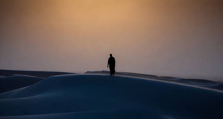 Silhouette d'une figure solitaire marchant le long des dunes du désert au coucher du soleil avec un ciel orange